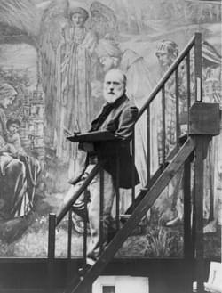 A black and white photo of Edward Burne-Jones. He has white hair and a beard. He's standing in front of one of his paintings.