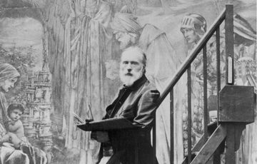 A black and white photo of Edward Burne-Jones. He has white hair and a beard. He's standing in front of one of his paintings.