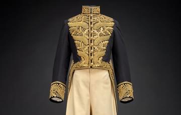 Two piece suit of gold-embroidered, high collared navy blue jacket with tails and light-coloured three-quarter length breeches