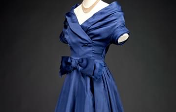 Mid-length dress made with shiny blue fabric, with short sleeves, a shawl collar and a large bow at the waist on the front in the same material