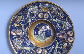 Round ceramic dish with a blue and white patterna around the main part, a circle in the middle has a painting of a cherub with wings