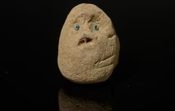 Small egg-shaped object formed of mud, two blue beads represent eyes and a mouth is moulded underneath to form a face