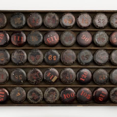 Rectangular wooden tray divided into five rows filled with round metal boxes, some have numbers painted on them in red