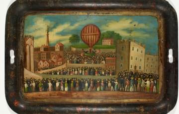 Metal traydecorated with a landscape painting showing crowds of people looking at a hot air balloon ascending