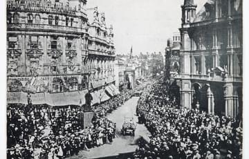 Black and white photograph of a city street full of people on the pavements, two cars drive down a road in the centre