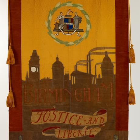 Large cloth banner with an appiled pattern in fabric of coat of arms surrounded by leaves above a silhouette of a city skyline with the words Birmingham and Justice and Liberty