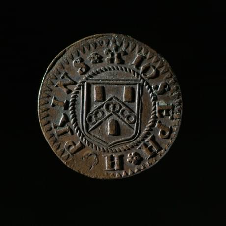 Dark coloured coin with coat of arms in the centre encircled by wording saying 'Joseph Perkins'