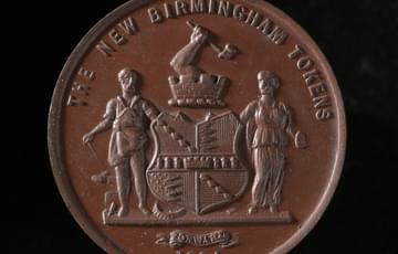 Brown coin with a coat of arms, two figures standing either side and an arm holding a hammer emerging from a turret above