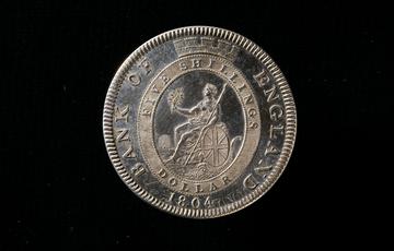 Silver coin featuring woman sitting down with a spear resting in one hand and a leafed branch in the other. A Union Jack shield rests at her side above a horn. At the top of the coin is a castle turret
