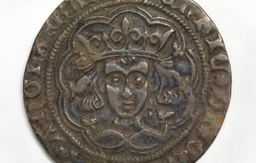 Dark coin with the head of a crowned, curly-haired figure looking straight ahead, inscribed