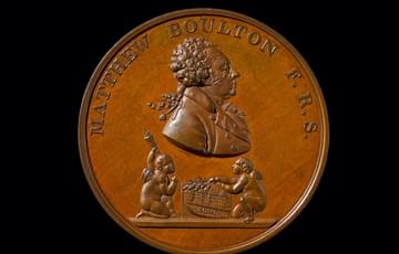 Brown coin featuring a man's bust in the centre and two cherubs below, one of which is holding a branch of leaves over a building