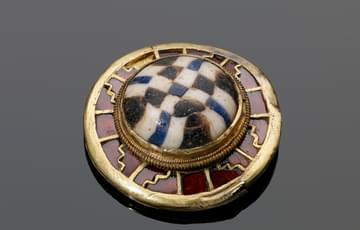 Small, round gold object set with garnet stones, in the centre is a raised black and white chequerboard pattern