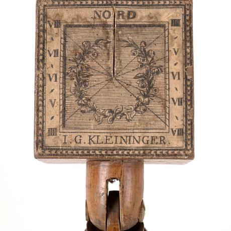 A square shaped object attached to a wooden handle. Roman numberals are painted either side and the word 'Nord' is painted at the top