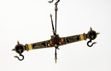 Thick gold and black beam with black hooks at either end. A long pole is attached to the centre of the beam perpendicularly. Either side of the pole are coats of arms in blue, red and gold