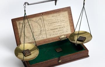 A wooden box, open and lined with green felt. Above it there is a beam from which two brass discs are suspended, balancing. There are small square objects in each disc