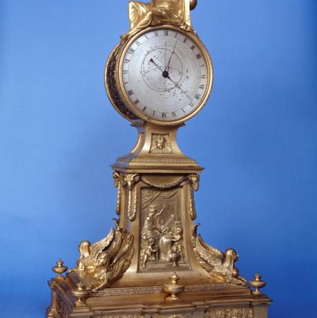 Gold coloured, ornate clock. A gold robed figure reclines on the top of the clock whilst at on the large, square foot of the clock, winged figures sit, pointing outwards. In the centre of the clock stand, more figures are inscribed, in the midst of an eventful scene