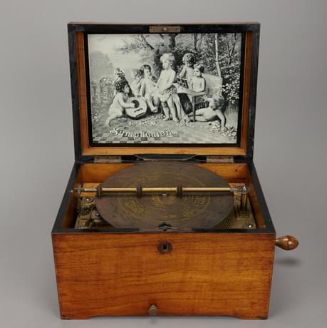 Open music box with a disc mechanism in the lower lid. In the upper lid is a black and white illustration of a group of partially-clothed children, surrounded by trees and nature. One plays a stringed instrument, another play the piano, and another appears to be singing. On the bottom left of the illustration is the word 'symphonion'.
