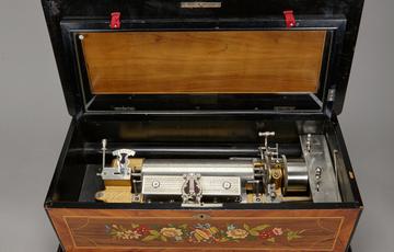 Open, rectangular music box with silver mechanisms sittting in the lower box. On the outside of the lower box is an illustration of flowers with a drum and brass instruments in the centre