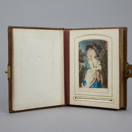 An object that looks like an open photograph album. On the page is an illustration of a lady in a cream and blue dress, wearing a black and blue, feathered hat. Below the illutsration is a horizontal line, etched into the page