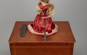 Figure of a monkey in a pink, satin dress and net head-piece, playing a stringed instrument, placed on top of a wooden box