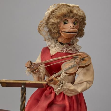 Close up of a figure of a monkey in a pink, satin dress and net head-piece, playing a stringed instrument and looking at sheet-music