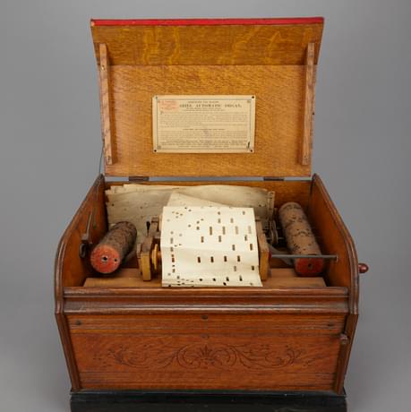 An open wooden box showing the internal workings of a music player, it contains cylinders with prongs and a paper roll with punched out squares