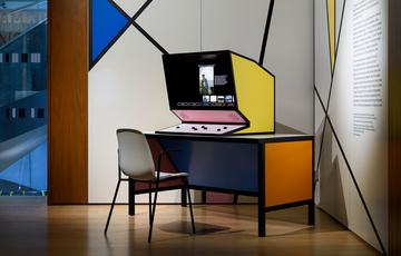 A computer in a yellow angular unit, sits on a orange and blue desk with a chair.