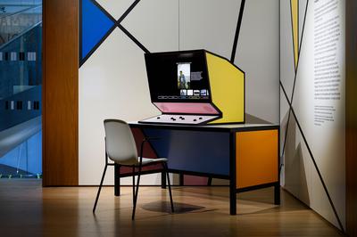 A computer in a yellow angular unit, sits on a orange and blue desk with a chair.