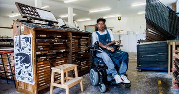 A man in an electric wheelchair sits in an room surrounded by art supplies and wooden draws and storage.