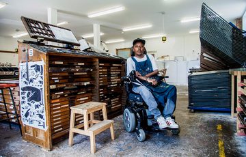 A man in an electric wheelchair sits in an room surrounded by art supplies and wooden draws and storage.