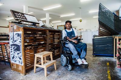 A man in an electric wheelchair sits in an room surrounded by art supplies and wooden draws and storage.