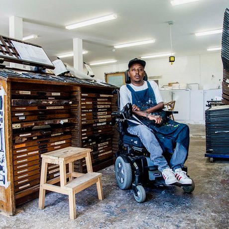 A man in an electric wheelchair sits in an room surrounded by art supplies and wooden draws and storage.