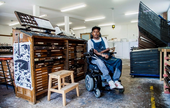 A man in an electric wheelchair sits in an room surrounded by art supplies and wooden draws and storage.