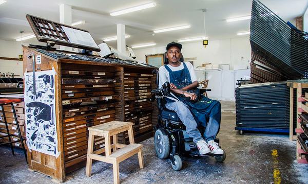 A man in an electric wheelchair sits in an room surrounded by art supplies and wooden draws and storage.