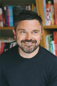 Portrait headshot of a man with black top and dark hair and beard, with a book case behind.