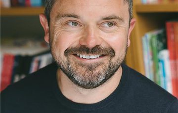 Portrait headshot of a man with black top and dark hair and beard, with a book case behind.