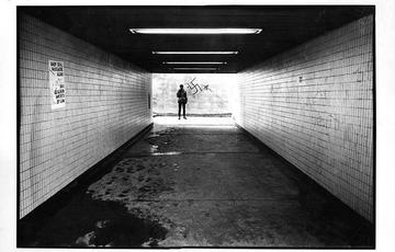 A person standing at the far end of an underpass, there is a swastika in graffiti behind the person and a there is a puddle of water on the floor in the foreground.