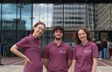 A group of three people wearing burgundy t-shirts that read 'Caramillo' standing outside a glass building.