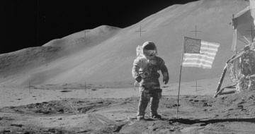 Black and white photo of an astronaut on the moon with USA flag next to him.