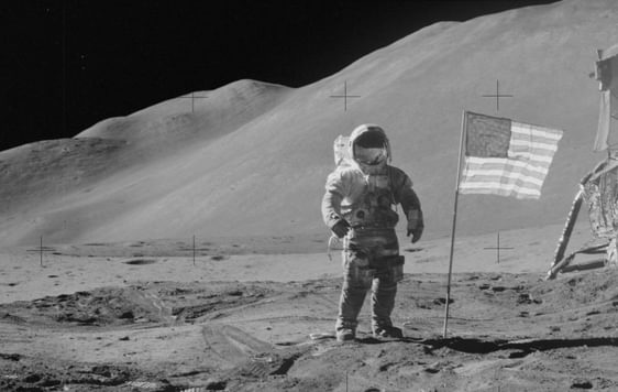 Black and white photo of an astronaut on the moon with USA flag next to him.