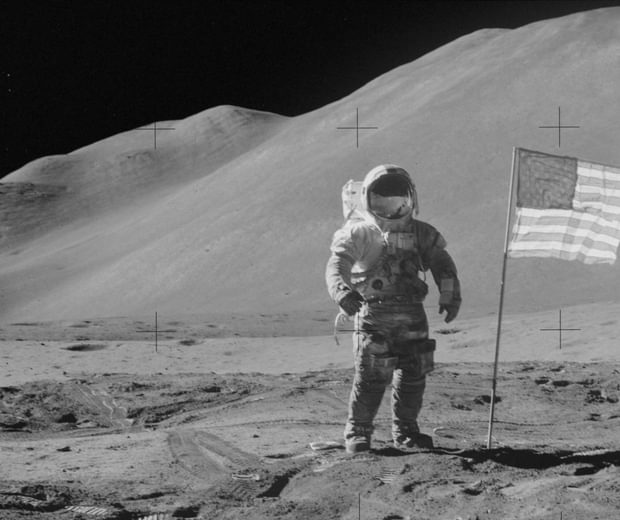 Black and white photo of an astronaut on the moon with USA flag next to him.