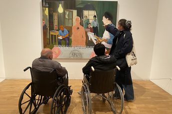 A person stands and points to an artwork on the while two people in wheelchairs look on.