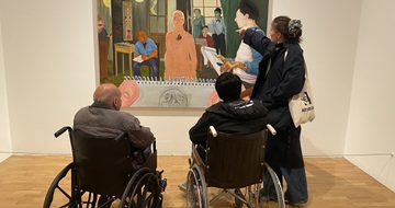 A person stands and points to an artwork on the while two people in wheelchairs look on.