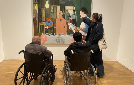 A person stands and points to an artwork on the while two people in wheelchairs look on.