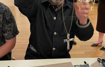 A person holding a necklace with a cross, they are seated at a table with craft supplies on it.