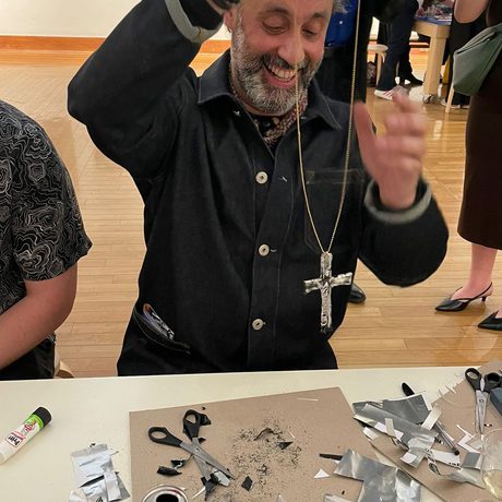 A person holding a necklace with a cross, they are seated at a table with craft supplies on it.