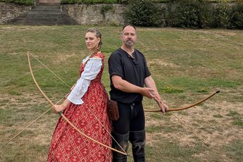 Two people pose back to back, they are dressed in historical costume and holding bows and arrows.