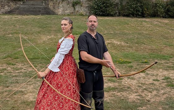 Two people pose back to back, they are dressed in historical costume and holding bows and arrows.