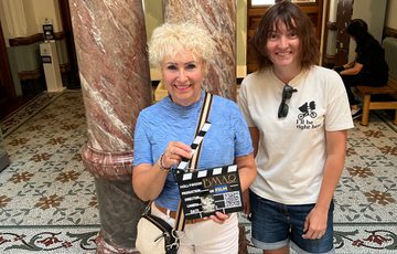 Two people inside a museum, one is holding a small clapperboard.