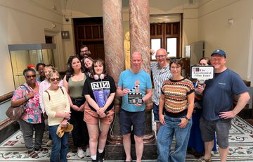 A group of people inside a museum, one is holding a small clapperboard.